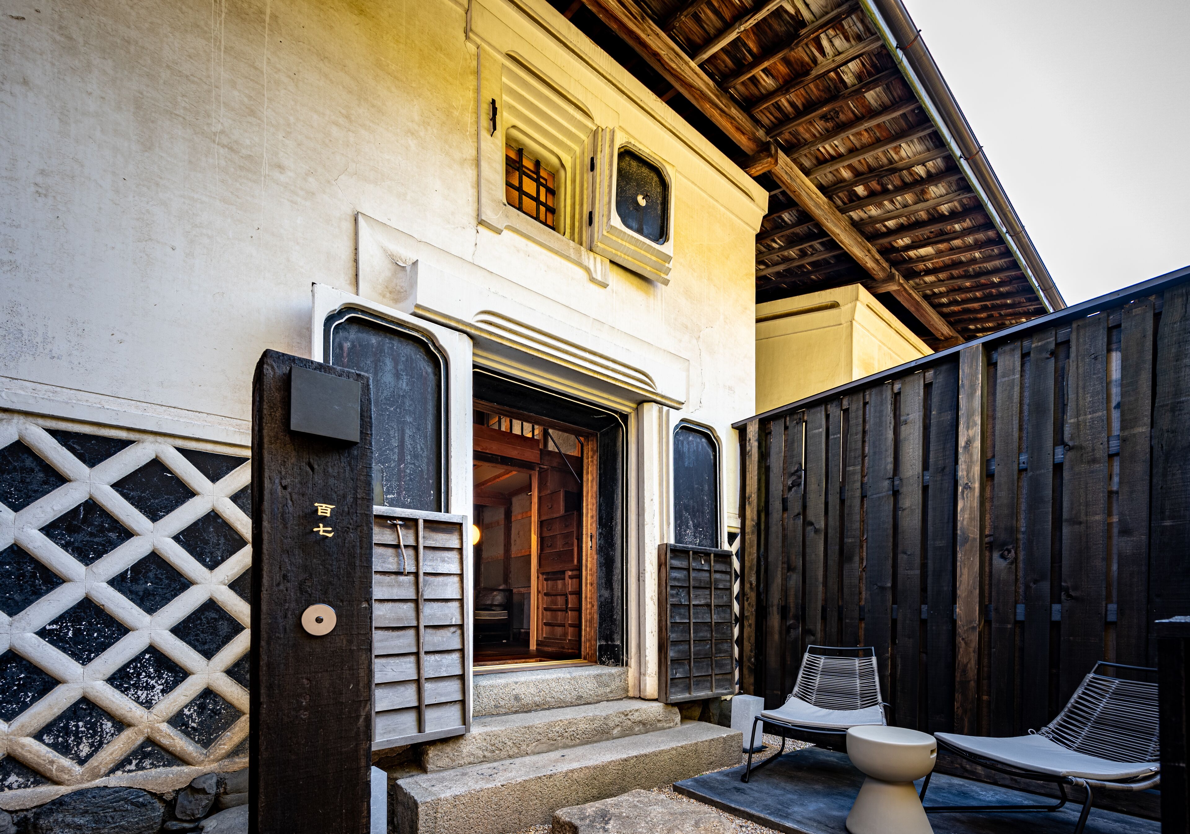A traditional building with a wooden door, steps leading to a porch, and two lounge chairs outside.
