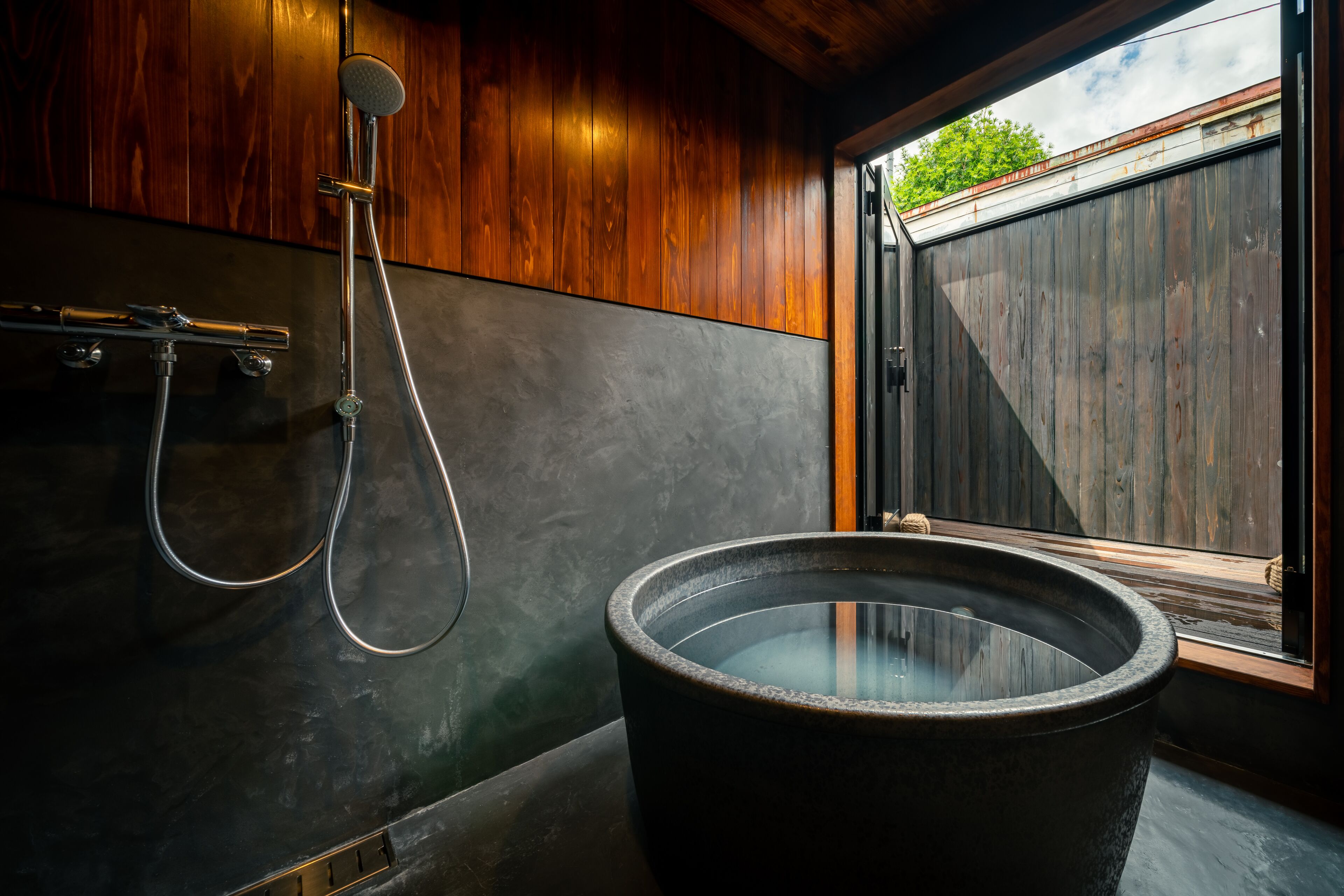 A modern bathroom with a large stone bathtub, a shower with a handheld showerhead, and a view of the outdoors through a wooden door.