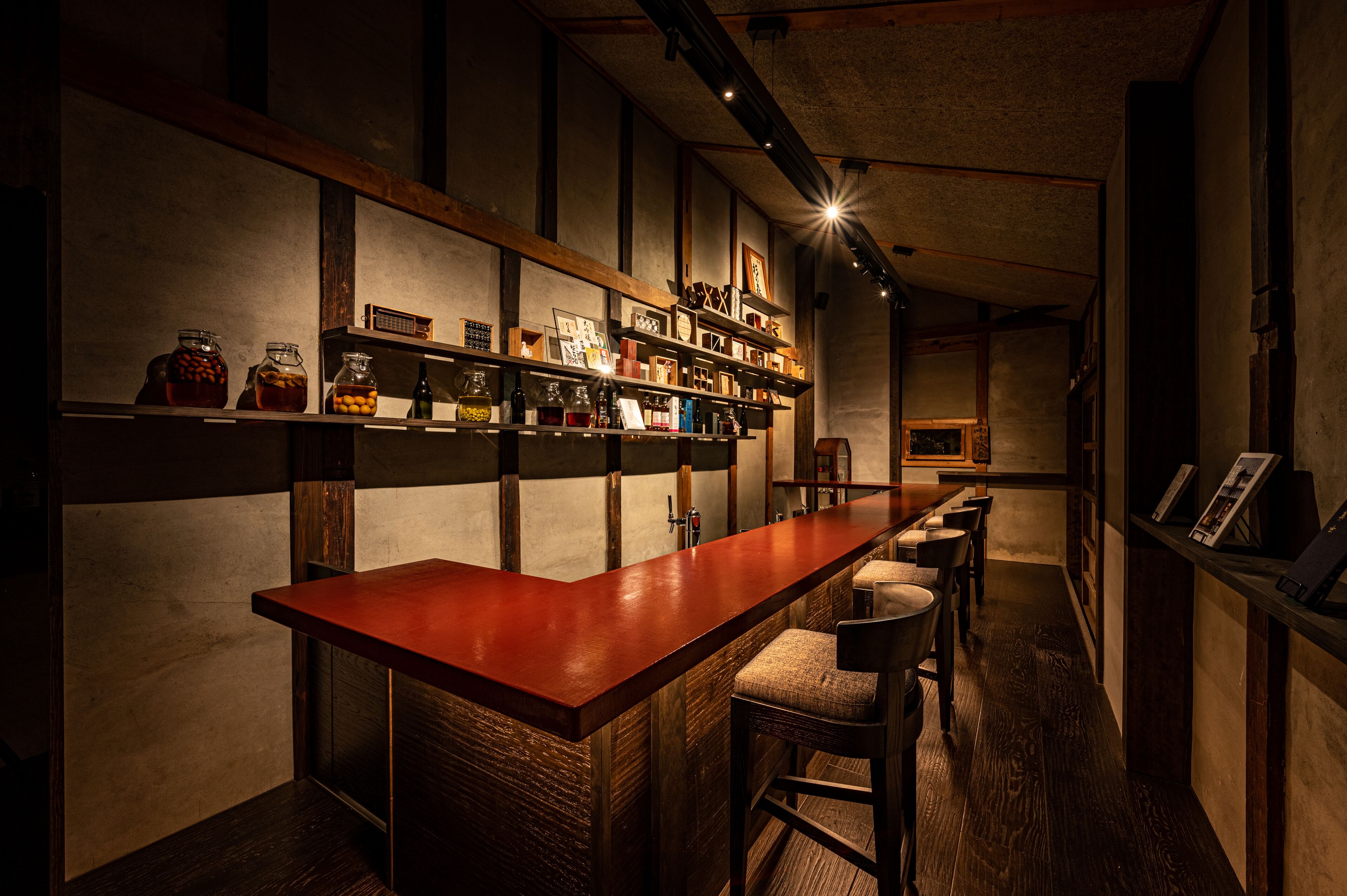 A bar area with a long counter, shelves with various bottles, and a few stools.