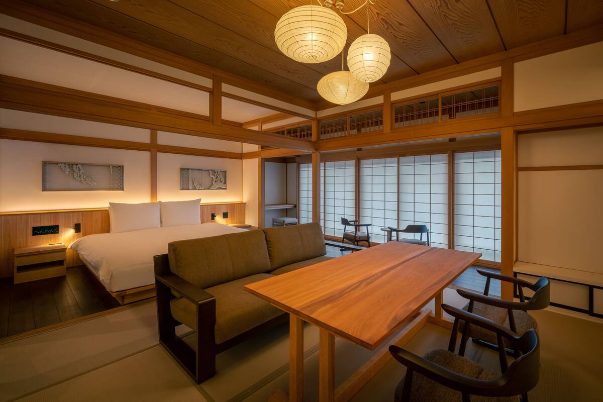 A traditional Japanese-style room with a wooden ceiling, tatami flooring, a low wooden table with chairs, a sofa, and a bed with white bedding.