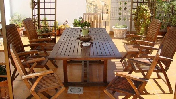 Una mesa de madera con sillas dispuestas a su alrededor, rodeada de plantas en macetas y un suelo de baldosas.