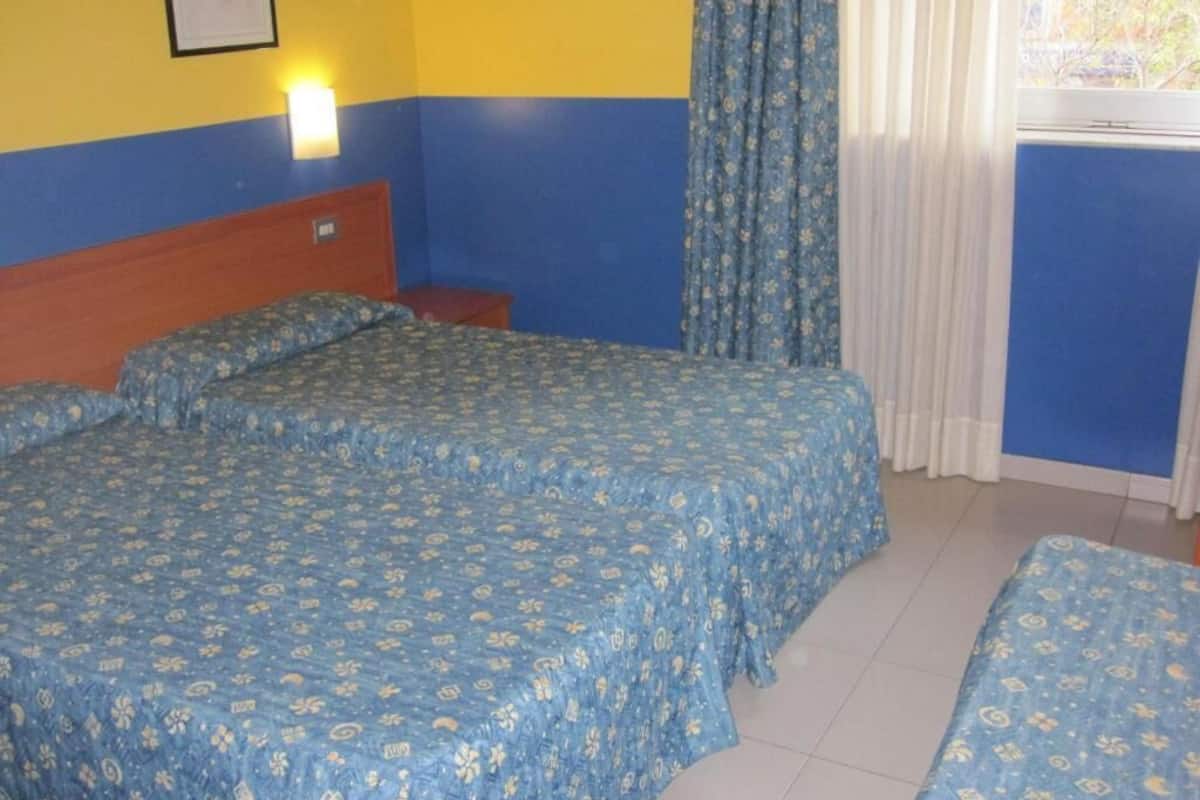 Una habitación con dos camas, una mesita de noche, una lámpara de pared y un ventanal con cortinas.