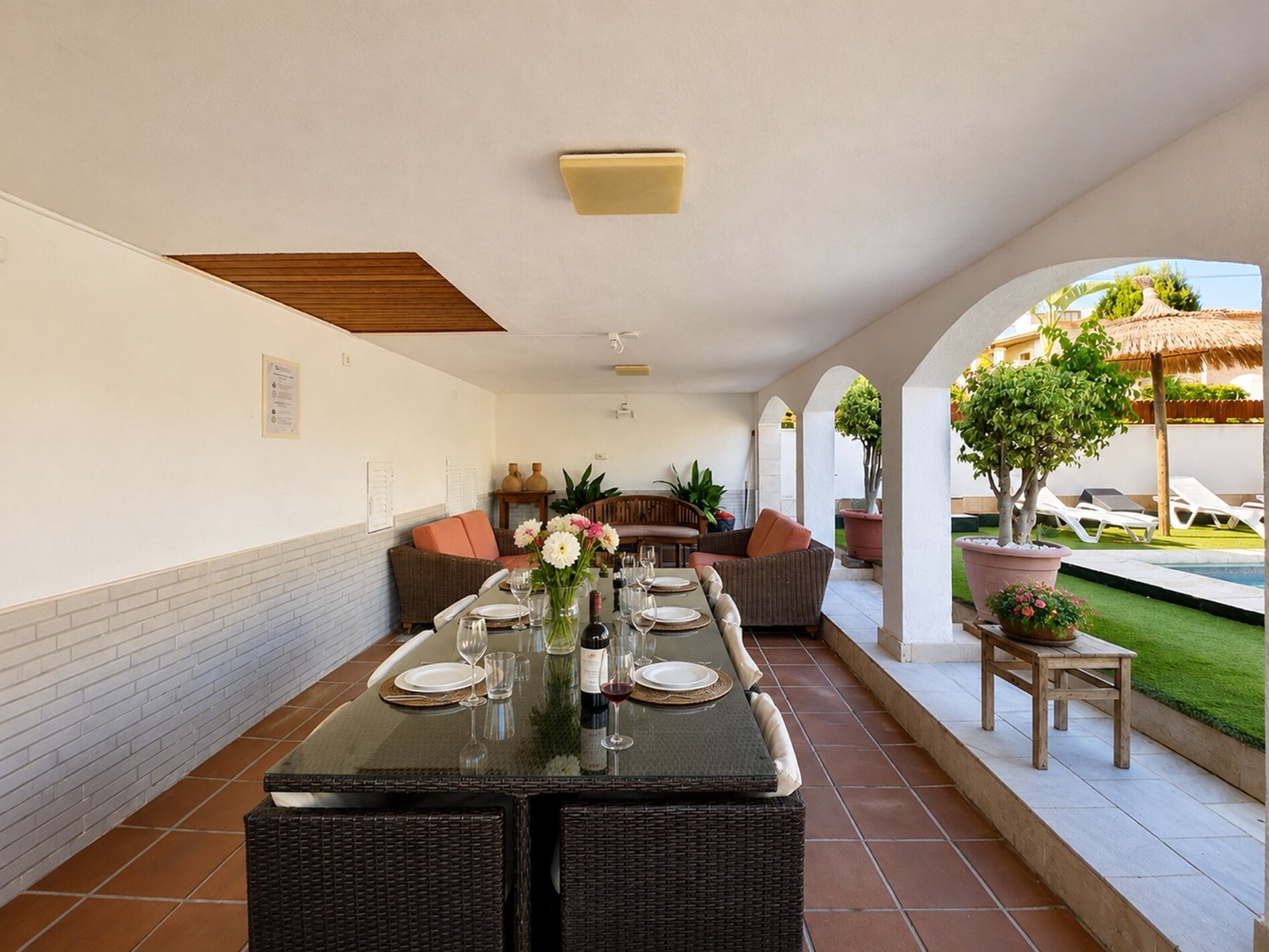 A dining area with a long table set for a meal, surrounded by wicker furniture and potted plants.