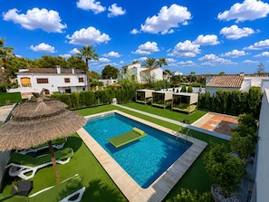 A swimming pool with a sun lounger and a thatched umbrella, surrounded by a well-maintained lawn and a variety of trees.