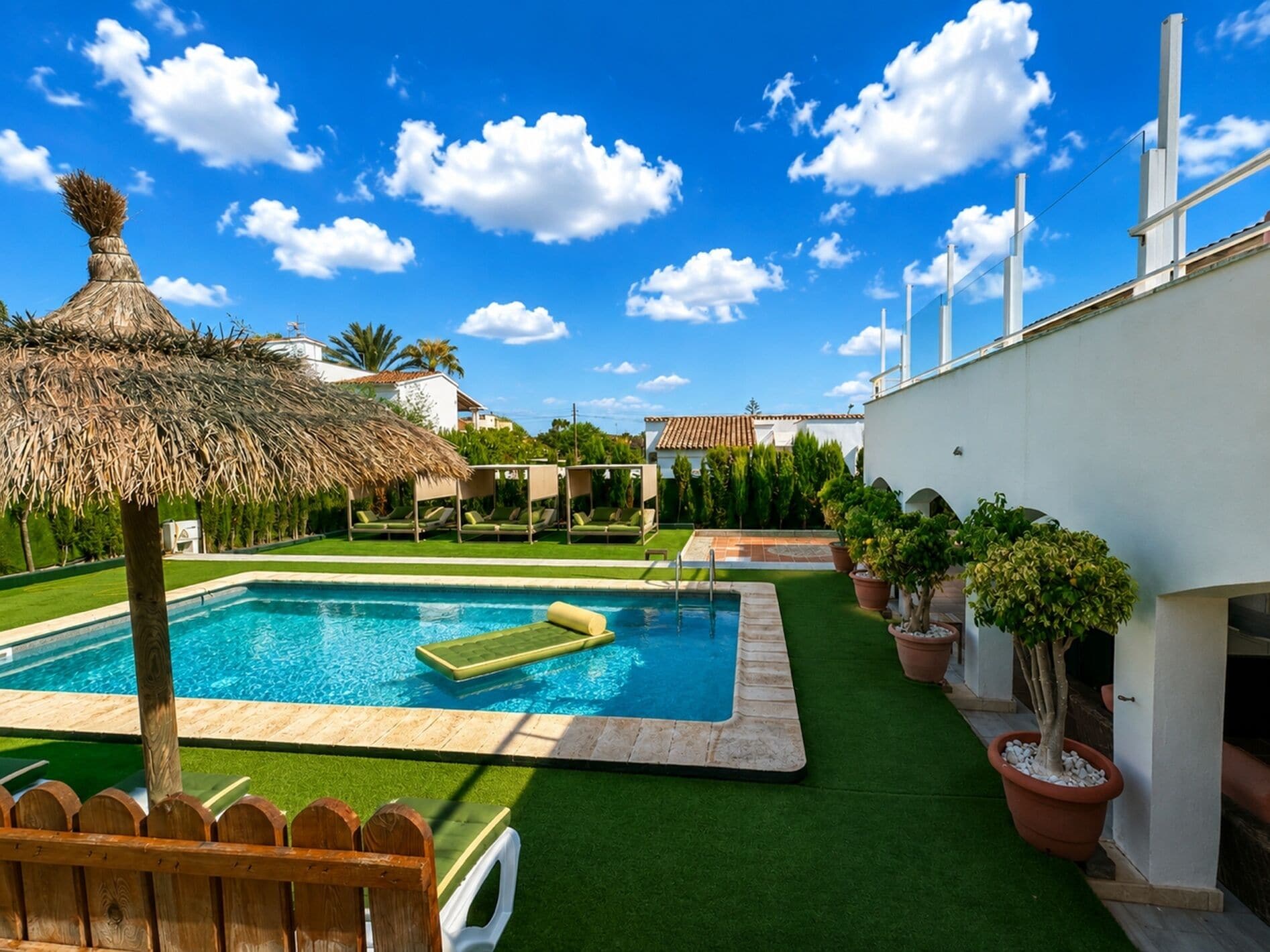 Ein Pool mit strohgedeckter Überdachung, umgeben von grünem Kunstrasen, Topfpflanzen und einem Holenzaun.
