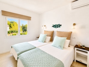 Cama doble con dos almohadas, mesita de noche, lámpara de pared y ventana con vistas a zonas verdes.
