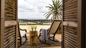 An open wooden door leading to a balcony with two rocking chairs, a small wooden table with two green glasses, and a view of a palm tree and distant landscape. - León de Sineu - Ten Bedroom Villa, Sleeps 21 (Sineu)