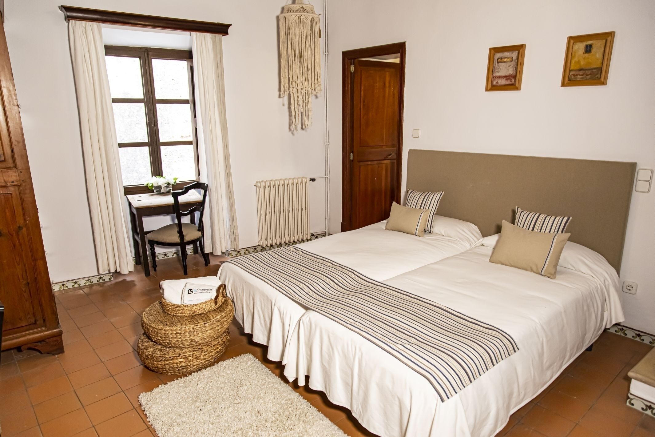A bedroom with a large bed, a wooden headboard, a desk with a chair, a radiator, and a window with curtains.
