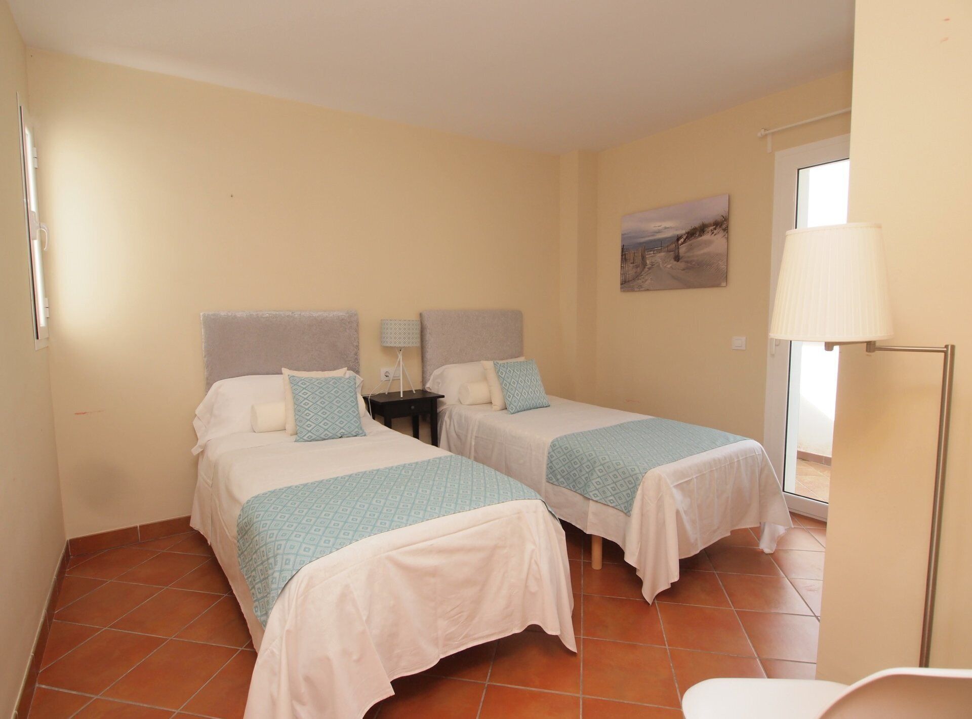 Una habitación con dos camas, cada una con cabecera, mesita de noche, lámpara y un cuadro en la pared.