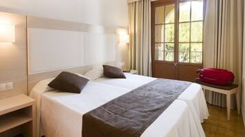 Una habitación de hotel con cama, mesitas de noche, una ventana con cortinas y una maleta roja sobre una pequeña mesa.
