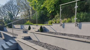 Un escalier extérieur moderne, avec des rampes en métal, entouré d’un jardin aménagé avec de petits arbustes et un mur en pierre.
