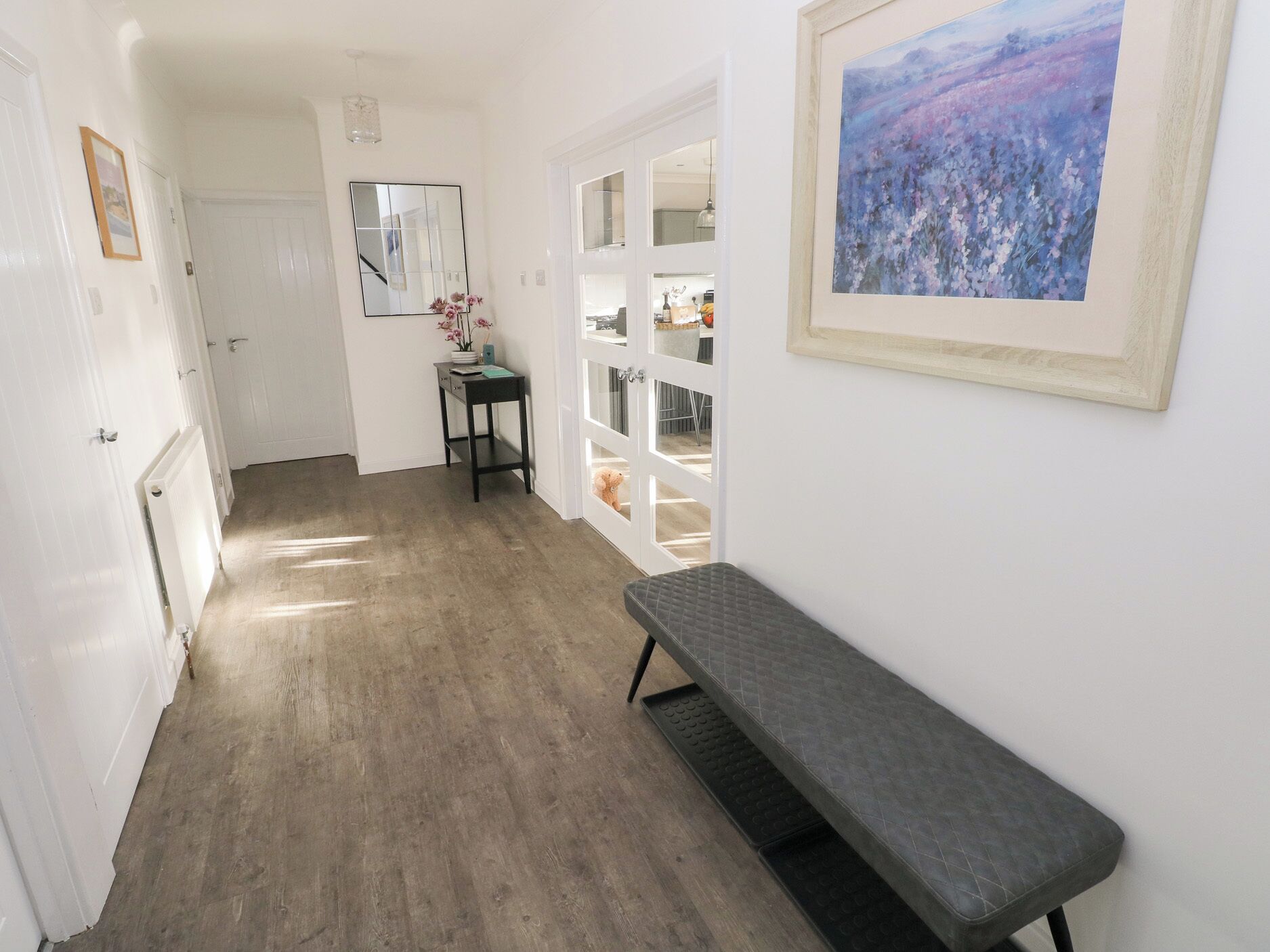 Käytävässä on penkki, peili, pieni pöytä, jossa on kukkia, ja seinällä on kehystetty taulu.