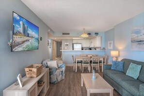Ein Wohnzimmer mit einer Couch, einem Couchtisch und einem Fernseher, der eine Strandansicht zeigt. Im Hintergrund ist eine Küchenzeile mit Esstisch und Stühlen zu sehen.
