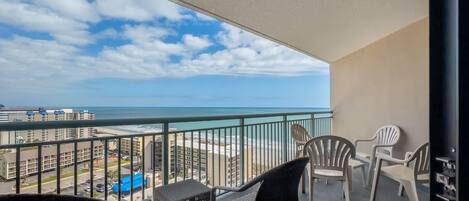 A balcony with wicker furniture, a glass railing, and a view of the ocean and cityscape.