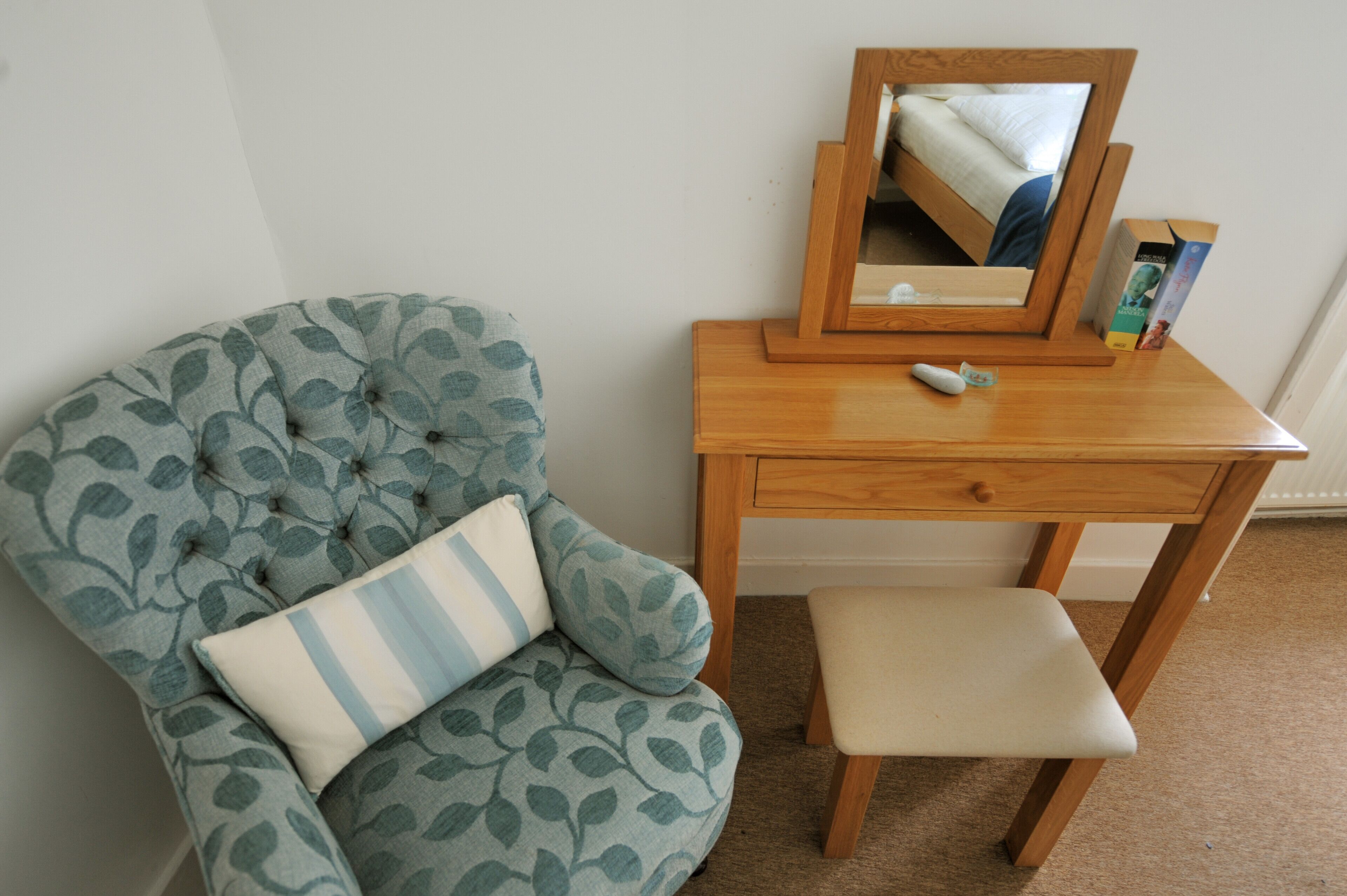 A room with a tufted armchair with leaf patterns, a wooden desk with a mirror, and a small stool.