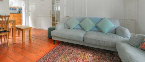 A living room with a sofa, a dining table, chairs, a patterned rug, and a kitchen area in the background.