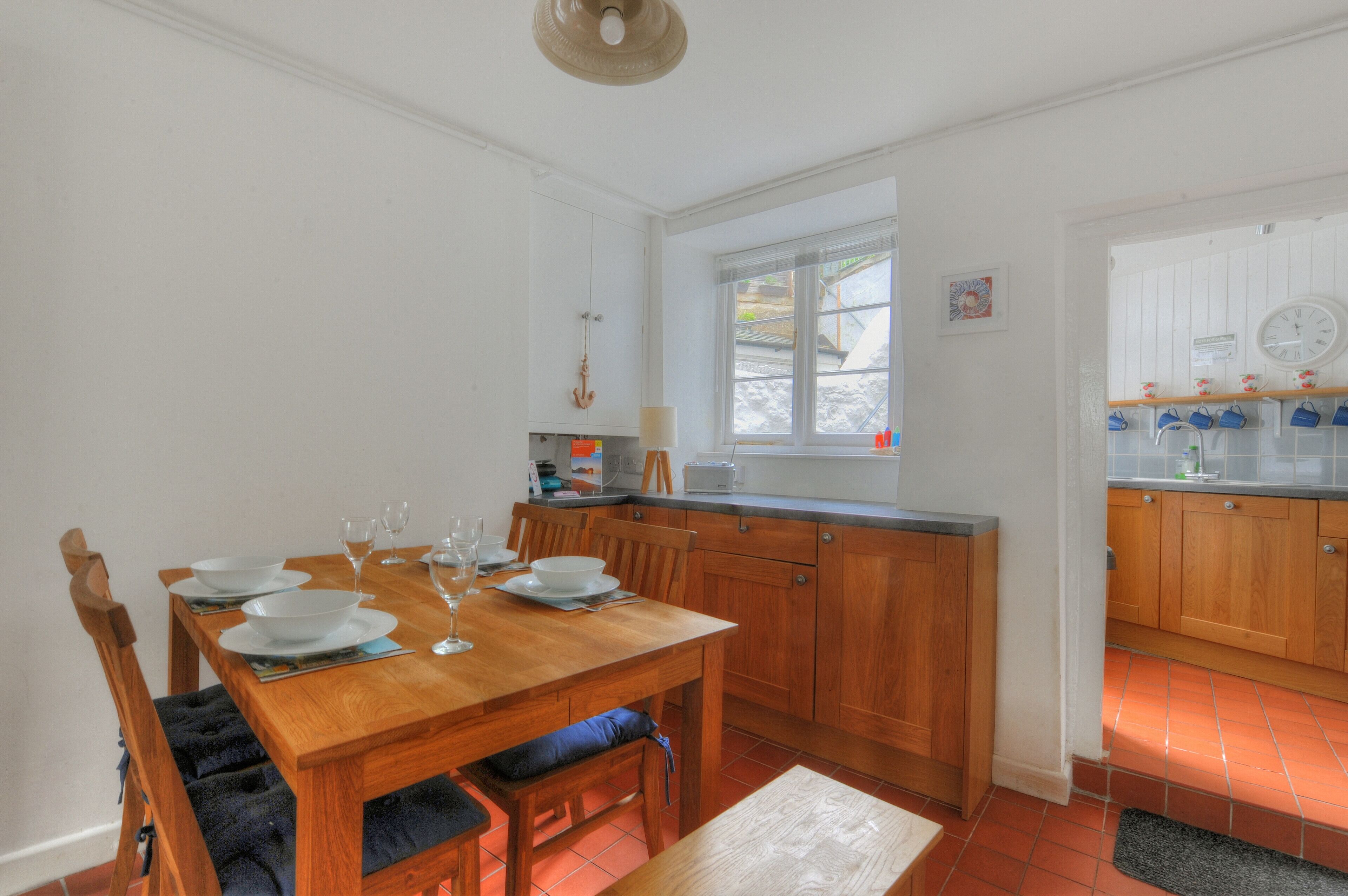 A dining area with a wooden table set for four, a window with curtains, and a kitchen with wooden cabinets and a tiled backsplash.