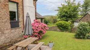 A wooden picnic table with benches, a grey umbrella, and pink flowers in a garden with stone walls and a grassy lawn. - Ash Cottage (Ilfracombe)