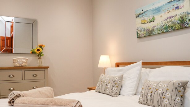 A neatly made bed with white linens and floral-patterned pillows, a bedside lamp, and a painting of a beach scene on the wall. - Ash Cottage (Ilfracombe)