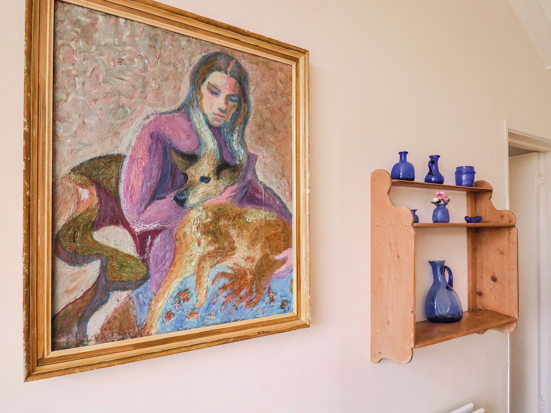 Un cuadro muestra a una mujer y un perro en la pared, debajo hay un estante de madera con jarrones azules.