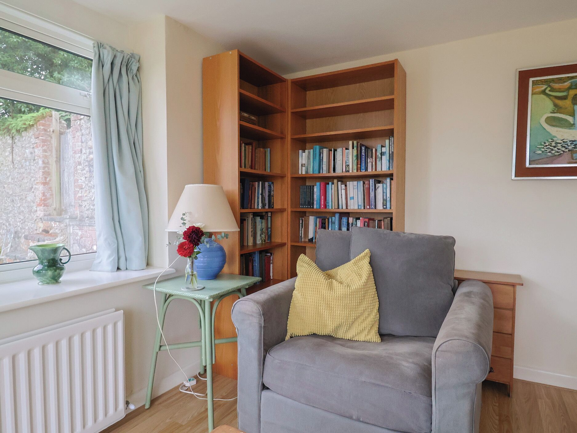 A cozy living room with a grey armchair, a small table with a lamp and flowers, a bookshelf, and a radiator.