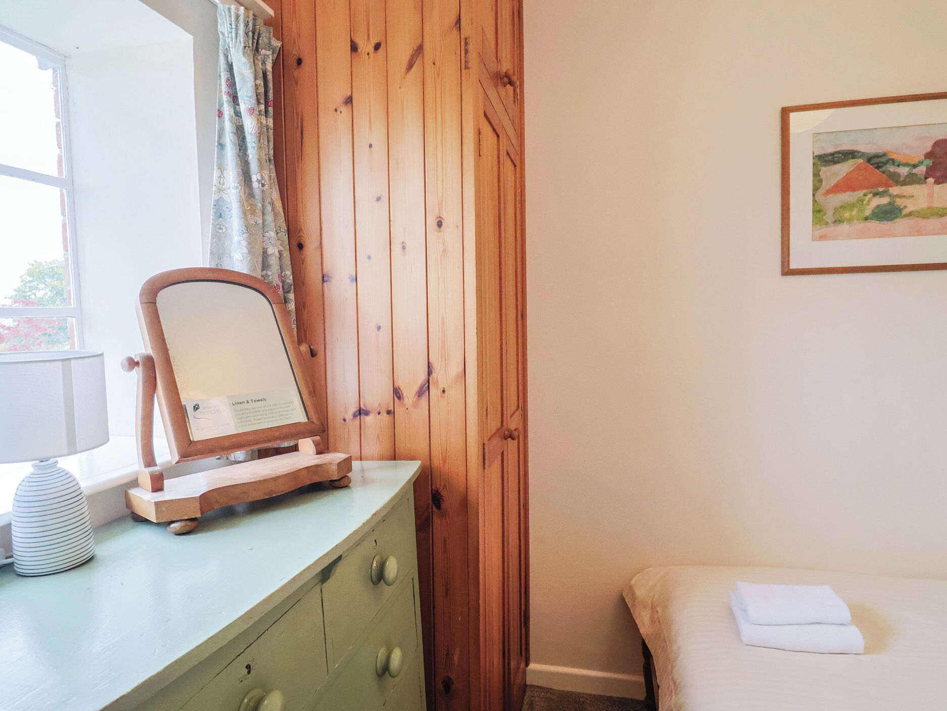 A room with a wooden wardrobe, a mirror on a dresser, a bed with a white pillow, and a painting on the wall.