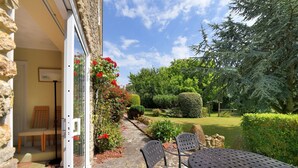 An outdoor patio with a table and chairs, a stone pathway, and a garden with various plants and a hedge. - Manor Lodge (Bridport)