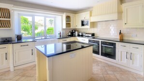 A modern kitchen with white cabinetry, a central island, and a large window providing a view of greenery outside. - Manor Lodge (Bridport)