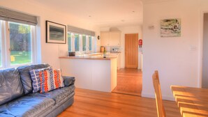 A modern living room with a leather sofa, a wooden dining table, and a kitchen area with white cabinets. - Pendower (Bridport)