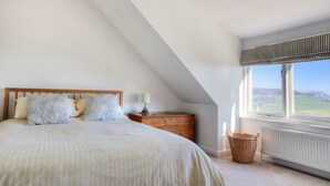 A bedroom with a sloped ceiling, a bed with patterned pillows, a wooden dresser, a lamp, a wicker basket, and a view of the countryside through a window. - Pendower (Bridport)