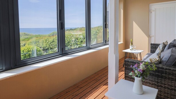 Un balcone con vista sul mare, un divano in vimini, un tavolino con un vaso di fiori e una bottiglia di vino.