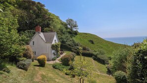 A white house with a red chimney, surrounded by lush greenery and a clear view of the sea. - Lynch Cottage (Lyme Regis)