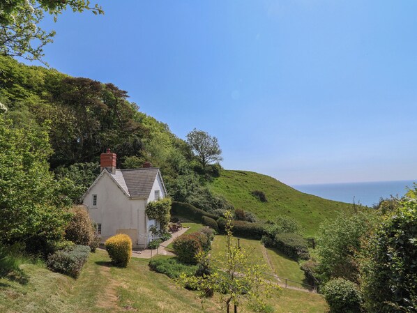 A white house with a red chimney, surrounded by lush greenery and a clear view of the sea.