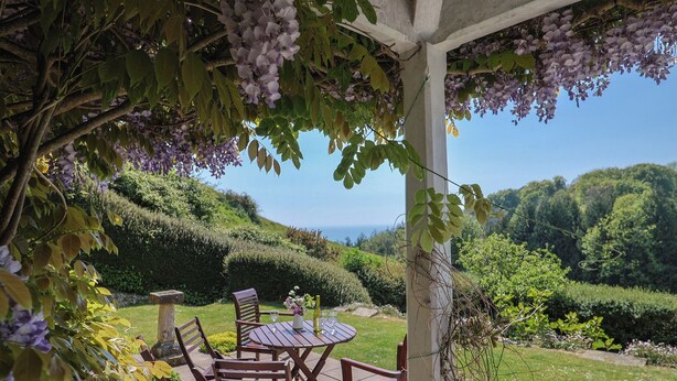 A covered outdoor seating area with a table and chairs, surrounded by lush greenery and a view of rolling hills. - Lynch Cottage (Lyme Regis)