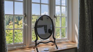 A room with a large window, a round mirror on a stand, and a view of greenery outside. - Lynch Cottage (Lyme Regis)