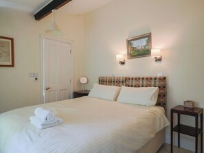 A neatly made bed with white linens and a rolled towel, a plaid headboard, a framed picture on the wall, and a bedside table with a lamp.