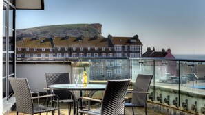 Een balkon met rieten stoelen en een tafel, met uitzicht op een kustplaatsje met terrassen en een klif.