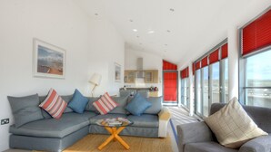 Een moderne woonkamer met een grijze bank, een salontafel en een grijze fauteuil. Achtergrond toont een keuken met rode jaloezieën voor de ramen.