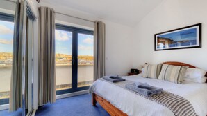 Een slaapkamer met een groot bed, een nachtkastje, een ingelijst schilderij aan de muur en uitzicht op de stad door het raam.