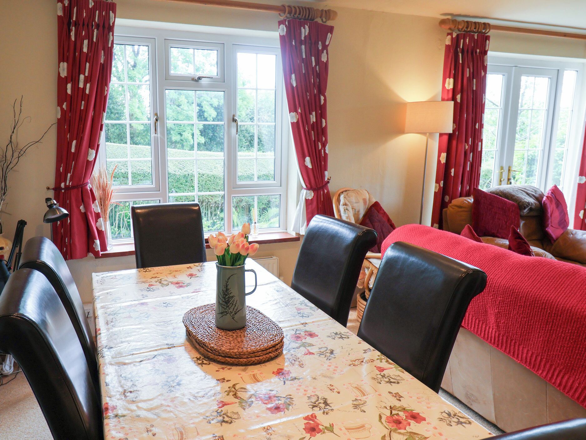A dining area with a table set for four, a vase of flowers, and a view of greenery through large windows.