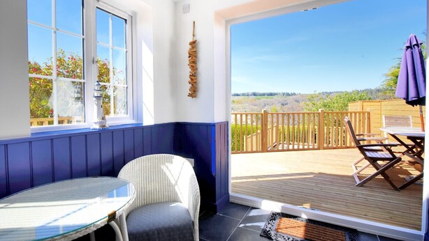 A room with a view of a wooden deck and a garden, a wicker chair, a small table, and a window with a view of trees. - Hillside Cottage (Lyme Regis)