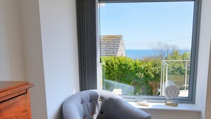 Ein Blick in ein Zimmer: ein grauer Sessel, ein Holzschrank und ein Fenster mit Ausblick auf Grünflächen und ein Gebäude.