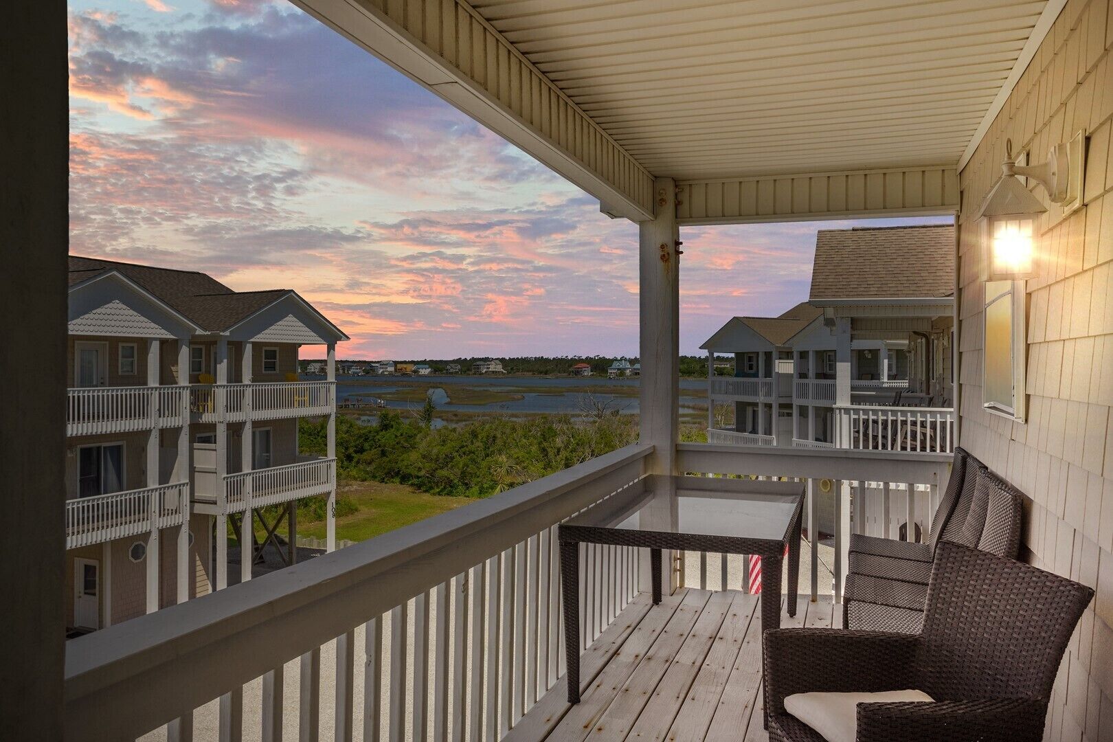 A covered balcony with wicker furniture, overlooking a residential area with multiple houses and a body of water.