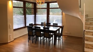 A dining area with a wooden table and chairs, large windows with a view of trees, and a unique staircase design. - Villa Golf (Karlovy Vary)
