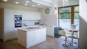 A modern kitchen with white cabinetry, a central island, and a dining area with bar stools. - Villa Golf (Karlovy Vary)