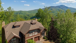 A two-story house with a sloped roof, a stone chimney, and a garage. It is surrounded by lush greenery and mountains in the background. - Bear Crossing at Echota - Long mountain views, indoor/outdoor pool access, hiking trails, gym (Banner Elk)