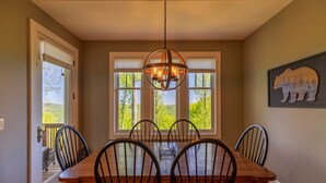 A dining room with a wooden table and chairs, a chandelier, and a large window with a view of trees. - Bear Crossing at Echota - Long mountain views, indoor/outdoor pool access, hiking trails, gym (Banner Elk)