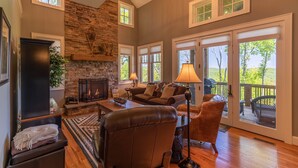 A spacious living room with a stone fireplace, comfortable seating, large windows, and a deck area. - Bear Crossing at Echota - Long mountain views, indoor/outdoor pool access, hiking trails, gym (Banner Elk)