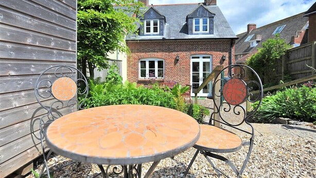 A patio with two round metal tables and chairs, a wooden fence, and a brick house with a gabled roof in the background. - 1 Woodroffe Meadow (Lyme Regis)