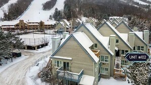 Un quartier résidentiel enneigé, avec plusieurs maisons, une piste de ski en arrière-plan et un panneau indiquant « UNITÉ N° 102 ».